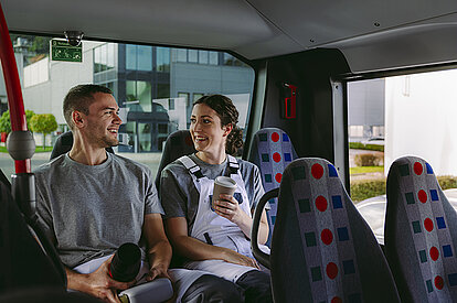 ein Mann und eine Frau mit Kaffee-togo-Bechern in der Hand sitzen im Bus und unterhalten sich