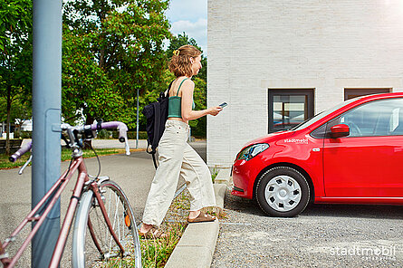 eine junge Frau mit Rucksack und Handy geht von ihrem abgestellten Fahrrad zu einem geparkten roten Cahrsharig-Fahrzeug von stadtmobil