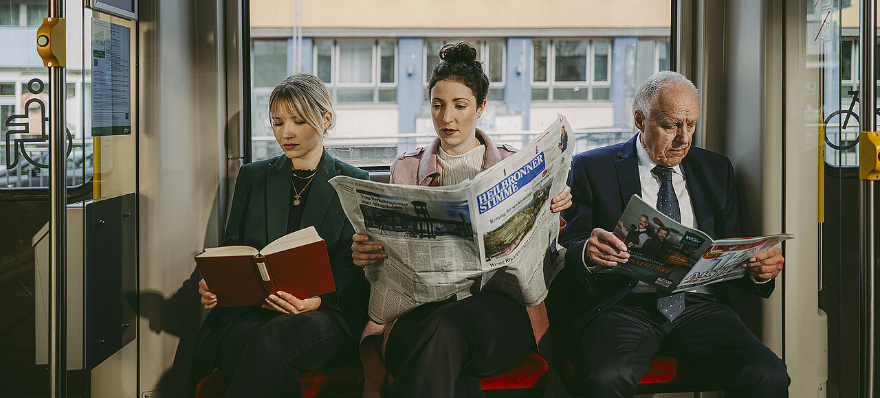 Zwei junge Frauen und ein älterer Herr sitzen in der Stadtbahn nebeneinander und lesen in unterschiedlichen Medien (Buch, Tageszeitung, Managermagazin)